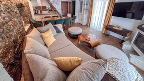 A living room with a sofa, ottoman, and a wooden coffee table. There is a dining area with chairs and a table, and a kitchen in the background.