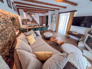 Een woonkamer met een bank, een voetenbankje en een houten salontafel. Er is een zithoek met stoelen en een tafel, en een keuken op de achtergrond.