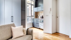 A compact kitchen with modern appliances, a sofa with a cushion, and a view of the outside through a window. - Blueground | Porte de Champerret, furnished (Paris)