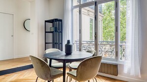 A room with a round table, two chairs, a black vase, a black bookshelf, and a large window with a view of trees and buildings. - Blueground | Porte de Champerret, furnished (Paris)