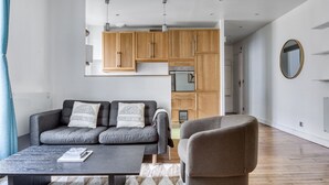 A modern living room with a grey sofa, a coffee table, and a brown armchair. There is a kitchen area with wooden cabinets and a built-in oven. - Blueground | Montparnasse, concierge, nr train (Paris)