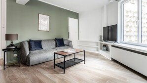 A modern living room with a grey sofa, a coffee table, a TV, and a large window with a view of a building. - Blueground | Villiers/Batignolles, nr shops (Paris)