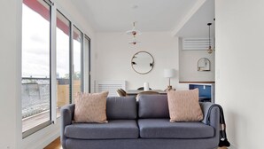 A modern living room with a grey sofa, two patterned cushions, a round mirror, and a dining area with a table and chairs. - Blueground | Chardon Lagache, balcony & concierge (Paris)
