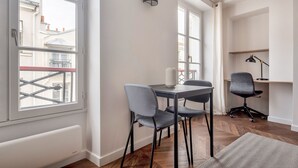 A modern, well-lit room with a dining table and chairs, a desk with a chair, and large windows offering views of the city.