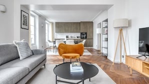 A modern living room with a sofa, a coffee table, and a chair. There is a kitchen in the background with cabinets and a dining area. - Blueground | Vendome/Saint Honoré, furnished (Paris)