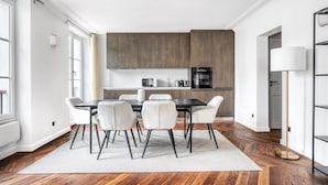 A modern kitchen with dark wooden cabinets, a black countertop, and a dining area with a black table and white chairs. - Blueground | Vendome/Saint Honoré, furnished (Paris)