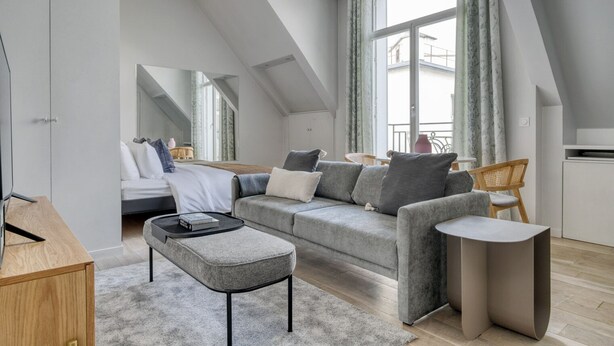 A modern loft bedroom with a large window, a grey sofa, a wooden desk, and a small dining table. - Blueground | George V, security, nr Champs Elysées (Paris)
