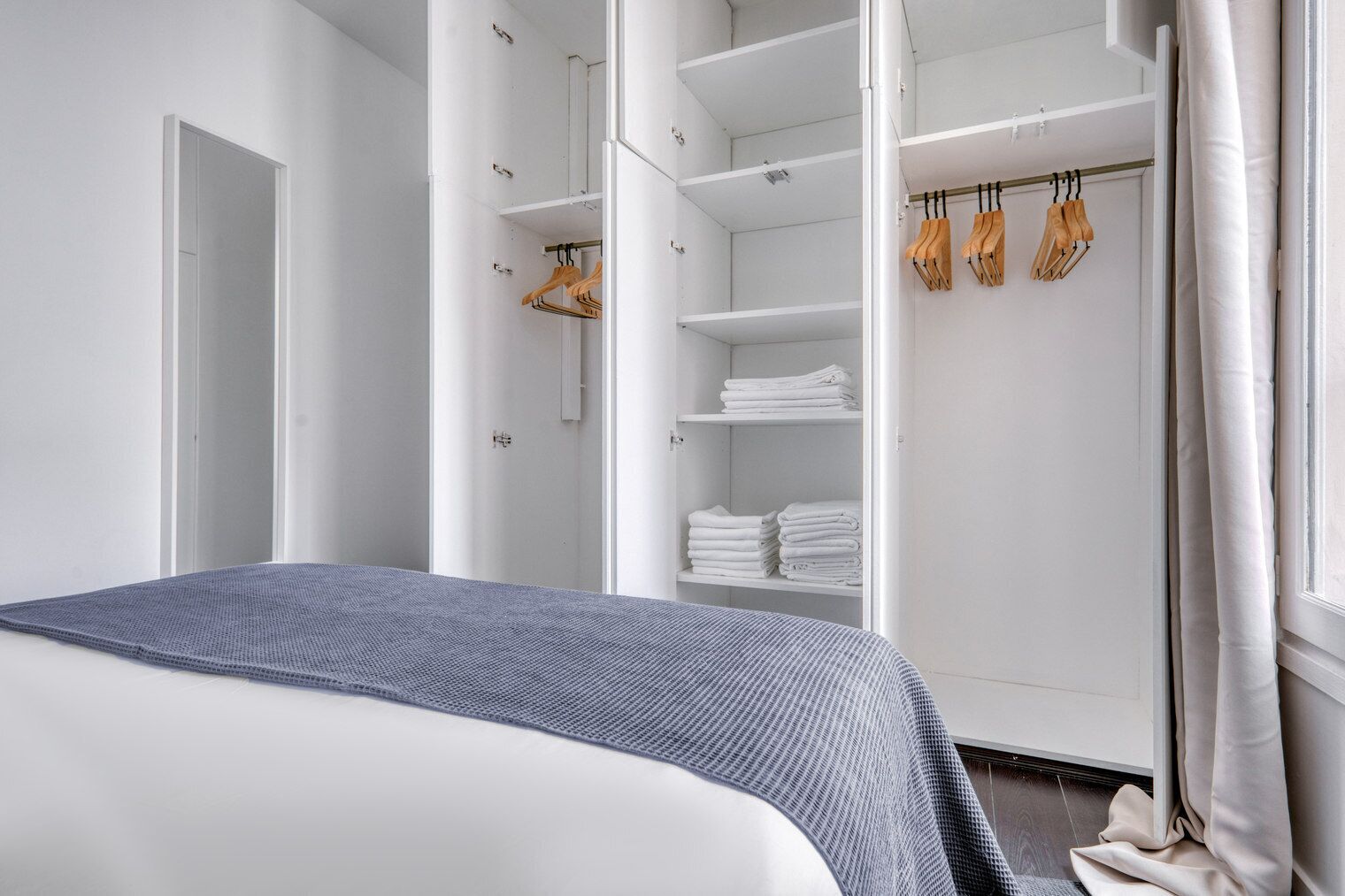 Ein modernes Schlafzimmer mit Bett, begehbarem Kleiderschrank inklusive Regalen und Ablagefläche sowie einem Fenster mit Vorhängen.