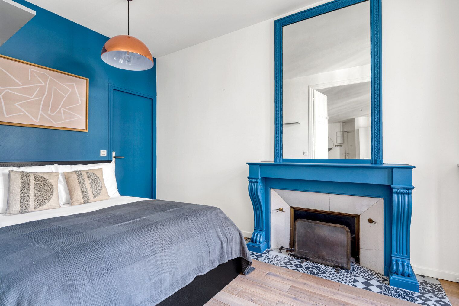 Un dormitorio con una cama, una chimenea azul, un espejo grande y una obra de arte mural geométrica.