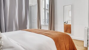 A bedroom with a bed, a window with curtains, a radiator, and a mirror on the wall. - Blueground | Batignolles, office & intercom (Paris)