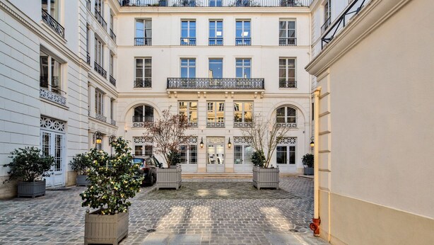 A courtyard with a building featuring a central entrance, multiple windows, and balconies. There are potted plants and a fire hydrant visible. - Blueground | Notre Dame de Lorette, nr opera (Paris)