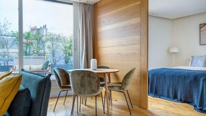 Et moderne hotelværelse med trægulv, et rundt bord med stole, en seng med blåt sengetæppe og et stort vindue med udsigt til bygninger og træer.