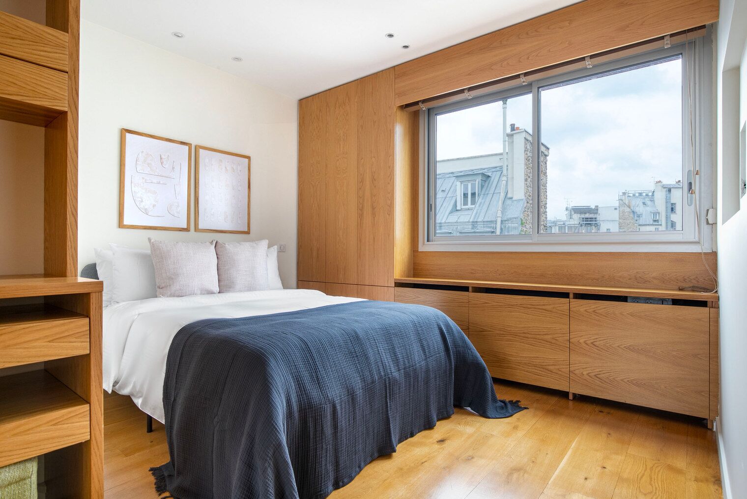 A bedroom with a bed, wooden wardrobe, and a large window with a city view.