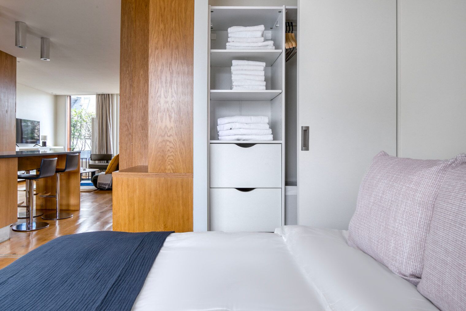 A modern hotel room with a large bed, a wooden accent wall, a built-in wardrobe with folded towels, and a dining area with bar stools.