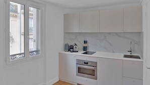 A compact kitchen with white cabinetry, a built-in oven, and a window with a view of buildings. - Blueground | Pigalle, furnished, nr opera (Paris)