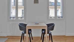 A room with a wooden table and four chairs, two windows with balconies, and a radiator. - Blueground | Pigalle, furnished, nr opera (Paris)