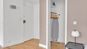 A room with a white door, a wooden floor, a small table with a lamp, and a closet with hanging clothes and an ironing board. - Blueground | Pigalle, furnished, nr opera (Paris)