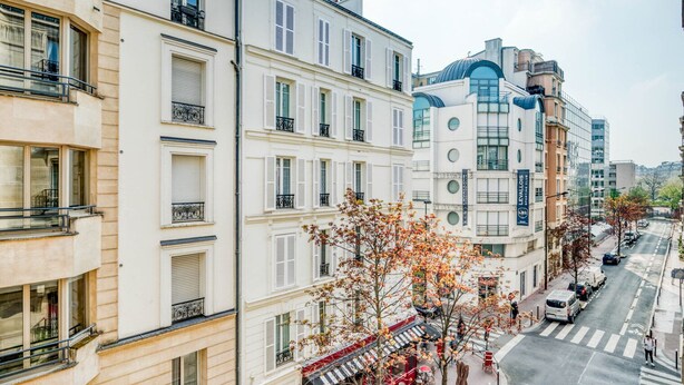 A row of multi-story buildings with balconies, a street with cars, and a café with outdoor seating. - Blueground | Levallois Perret, nr dining (Paris)