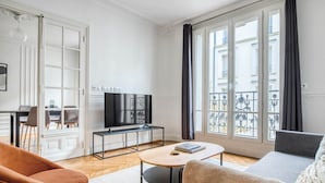 A modern living room with a sofa, a coffee table, a TV on a stand, and a dining area with chairs and a table. - Blueground | Levallois Perret, nr dining (Paris)