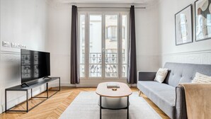 A modern living room with a flat-screen TV, a grey sofa, a coffee table, and a large window with curtains. - Blueground | Levallois Perret, nr dining (Paris)