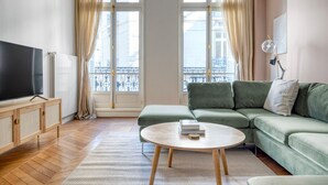 A living room with a green sofa, a round coffee table, a TV on a wooden cabinet, and large windows with curtains. - Blueground | Champs Elysées, furnished, balcony (Paris)