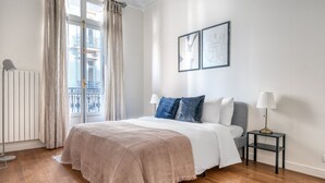 A bedroom with a bed, a bedside table with a lamp, a radiator, and a window with curtains. - Blueground | Champs Elysées, furnished, balcony (Paris)