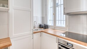 A modern kitchen with white cabinetry, a wooden countertop, a built-in oven, and a window with blinds. - Blueground | Champs Elysées, furnished, balcony (Paris)