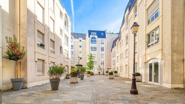 A narrow street with buildings on both sides, potted plants, and a clear sky. - Blueground | Le Marais, concierge & elevator (Paris)