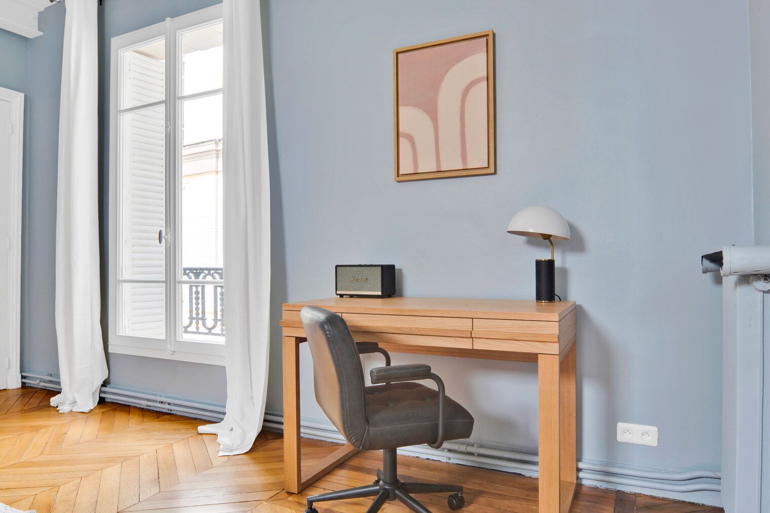 A room with a wooden desk, a chair, a lamp, a framed picture on the wall, and a window with white shutters.