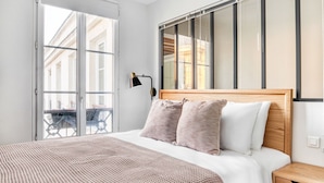A modern bedroom with a large bed, a wooden headboard, a nightstand, and a window with a view of buildings. - Blueground | St Paul, nr Bastille & Le Marais (Paris)
