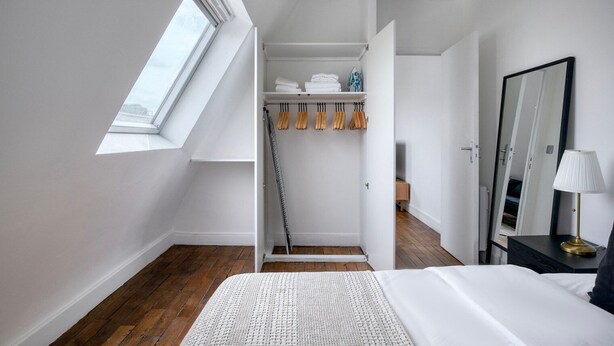Un dormitorio con tragaluz, cama, mesita de noche, espejo y armario con bolsos colgados.