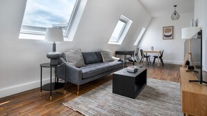 Un loft moderno con un sofá gris, una pequeña mesa redonda, una mesa baja negra y un escritorio de madera. Cuenta con tragaluces y un ventanal grande que proporciona luz natural.