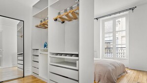 Une chambre moderne dotée d’une grande fenêtre, d’un lit recouvert d’une couverture tricotée et d’un dressing avec des étagères et un espace pour suspendre les vêtements.