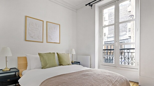 Une chambre à coucher comprenant un lit, deux tables de chevet, une fenêtre donnant sur des immeubles et deux tableaux encadrés accrochés au mur.