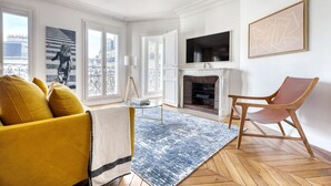 A living room with a yellow sofa, a blue rug, a fireplace, and a TV on the wall. - Blueground | Rochechouart, furnished, balcony (Paris)
