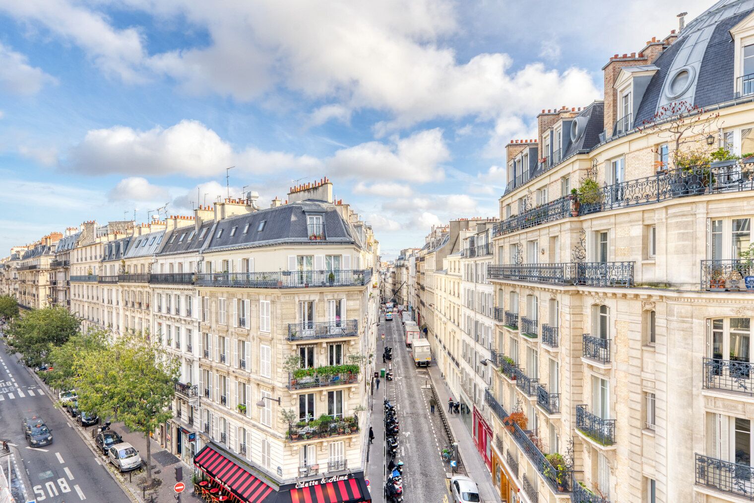 A street view of Parisian buildings with balconies, a café, and parked cars.
