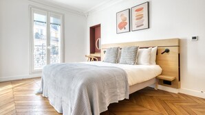 A bedroom with a bed, a wooden headboard, a bedside table, and a window with a view of the outside. - Blueground | Rochechouart, furnished, balcony (Paris)