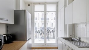 Cozinha compacta com geladeira, pia e janela com vista para uma varanda com guarda-corpo de metal.