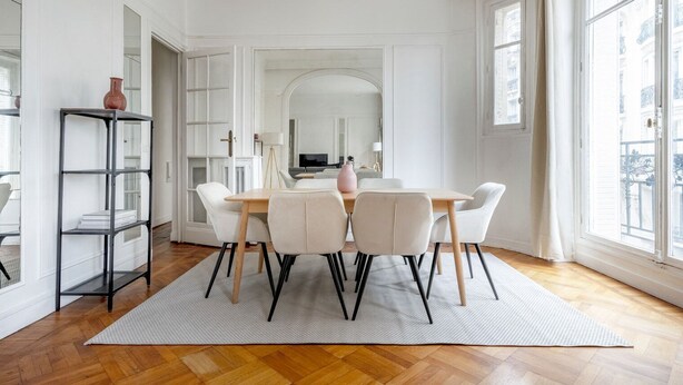Uma sala de jantar com uma mesa redonda, cadeiras brancas e um tapete. Há uma estante de livros e uma janela grande com vista para edifícios.