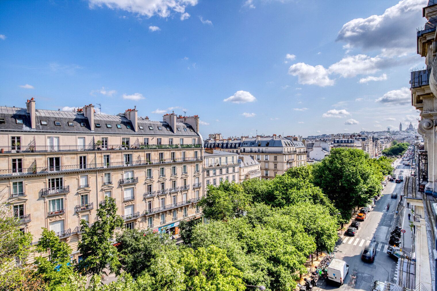 A row of classic Parisian buildings with balconies, a tree-lined street, and a clear blue sky.