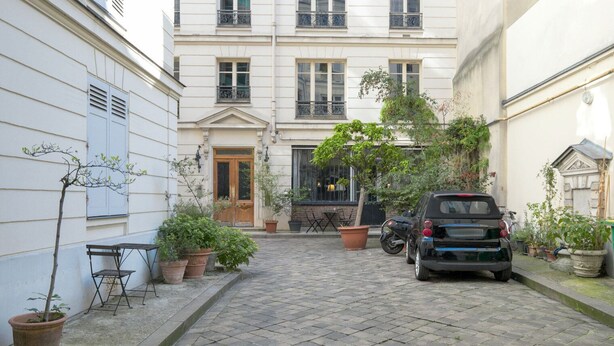 A narrow cobblestone alley with a parked car, potted plants, and a building with a wooden door and windows.