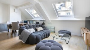 A modern loft apartment with a high ceiling, skylights, a grey sectional sofa, a glass coffee table, and a TV on a wooden cabinet.