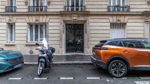 Une vue de rue avec une moto garée et une voiture devant un immeuble doté de détails architecturaux élaborés et d’un portail noir.