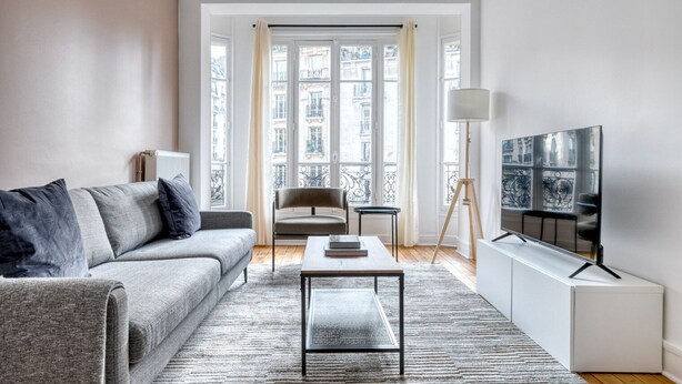 A modern living room with a grey sofa, a coffee table, a TV on a stand, and a view of buildings through large windows. - Blueground | Lamark-Caulaincourt, elevator (Paris)