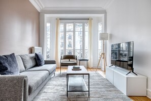 A modern living room with a grey sofa, a coffee table, a TV on a stand, and a view of buildings through large windows.