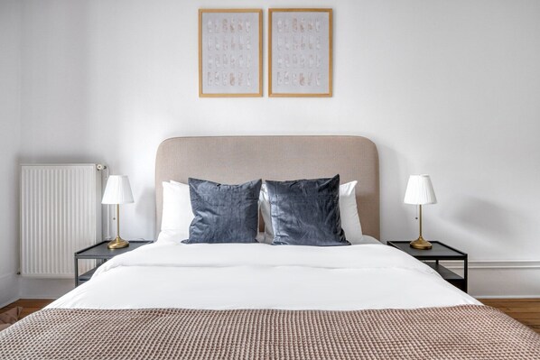A neatly made bed with a beige headboard, two bedside tables with lamps, and two framed artworks on the wall.