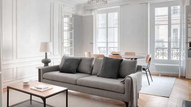 Ein modernes Wohnzimmer mit einem grauen Sofa, einem kleinen Tisch und einem Sessel. Große Fenster mit weißen Rahmen bieten einen Blick auf die Gebäude außerhalb.