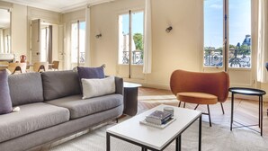 A spacious living room with a grey sofa, a small table with books, a red armchair, and large windows offering a view of the city. - Blueground | Invalides, balcony & elev nr gardens (Paris)