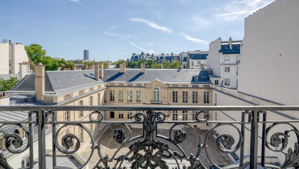 A balcony with ornate wrought iron railings overlooking a courtyard with a building featuring a tiled roof and a chimney. - Blueground | Invalides, balcony & elev nr gardens (Paris)
