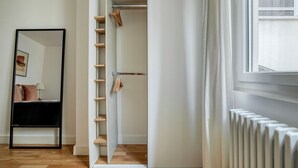 A hotel room with a wooden floor, a black-framed mirror, a white wardrobe with wooden shelves, and a white radiator. - Blueground | Jules-Joffrin, furnished & equipped (Paris)